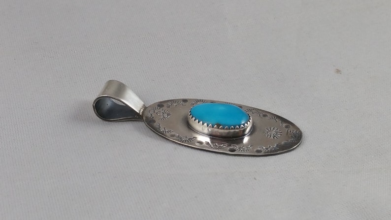 Pode incluir: Um pingente de prata com uma pedra turquesa ovalada engastada em uma moldura de prata. O pingente &eacute; decorado com um design de prata em torno da pedra e na parte de tr&aacute;s.