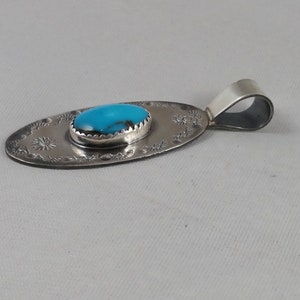 Pode incluir: Um pingente de prata com uma pedra turquesa engastada em forma oval. O pingente tem um design detalhado em torno da pedra e um la&ccedil;o para uma corrente.