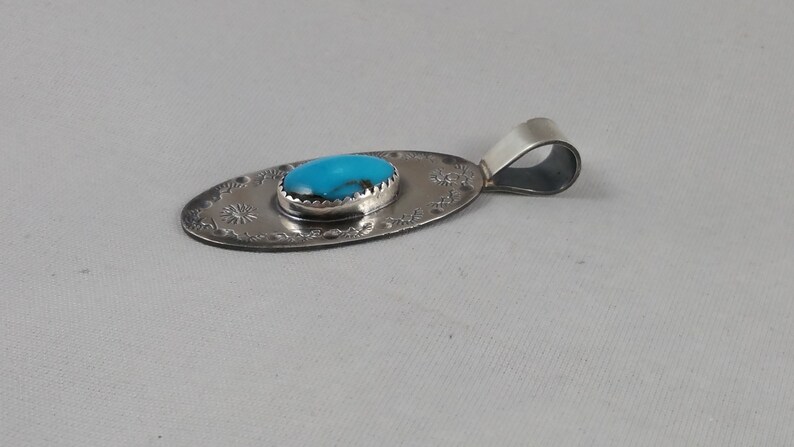 Pode incluir: Um pingente de prata com uma pedra turquesa engastada em forma oval. O pingente tem um design detalhado em torno da pedra e um la&ccedil;o para uma corrente.