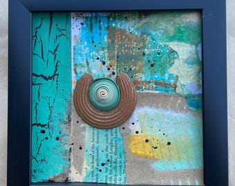 Puede incluir: Obra de arte abstracta de técnica mixta en un marco negro. La obra presenta una pieza metálica en forma de herradura con un centro en espiral. El fondo tiene pintura turquesa, azul y marrón con salpicaduras negras. La obra incluye texto de un libro.