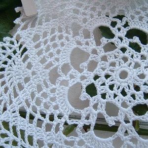 Peut inclure: Gros plan sur un napperon au crochet blanc fait &agrave; la main. Le motif de dentelle complexe pr&eacute;sente un motif floral central entour&eacute; de jours et de bords festonn&eacute;s. Le napperon est en fil blanc.
