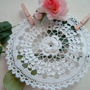 Puede incluir: Doily de ganchillo blanco con un delicado dise&ntilde;o floral en el centro. El doily se muestra con una rosa rosa y hojas verdes.