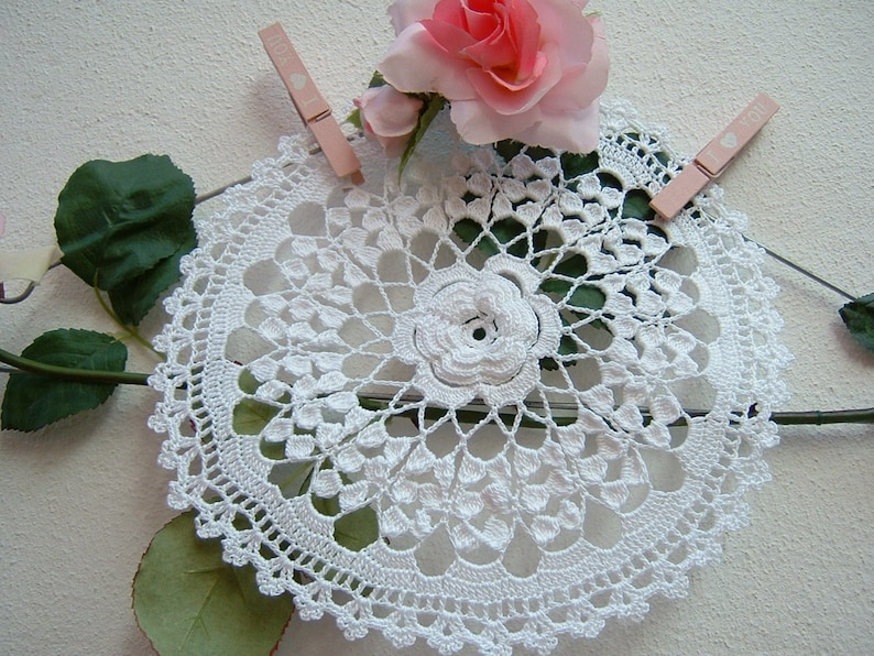 Puede incluir: Doily de ganchillo blanco con un delicado dise&ntilde;o floral en el centro. El doily se muestra con una rosa rosa y hojas verdes.