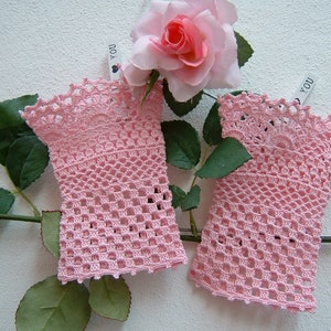 May include: A pair of pale pink, open-weave crochet fingerless gloves. The gloves have a scalloped edge and are displayed with a pink rose and green leaves. The gloves are a delicate, handmade accessory.