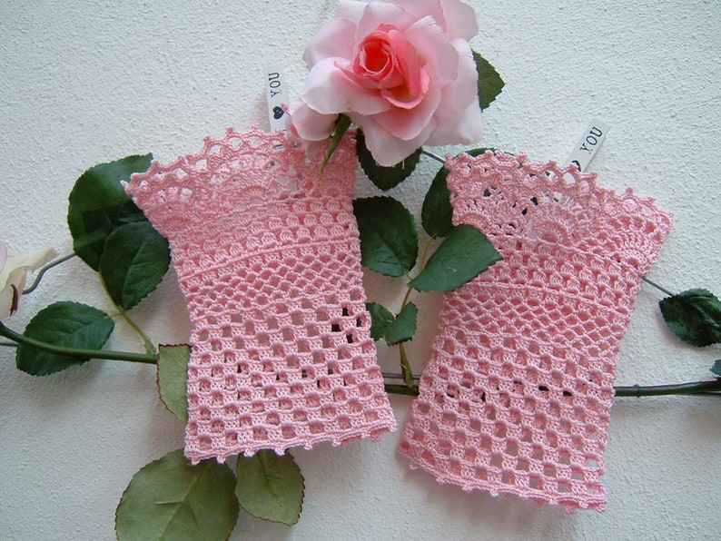 May include: A pair of pale pink, open-weave crochet fingerless gloves. The gloves have a scalloped edge and are displayed with a pink rose and green leaves. The gloves are a delicate, handmade accessory.