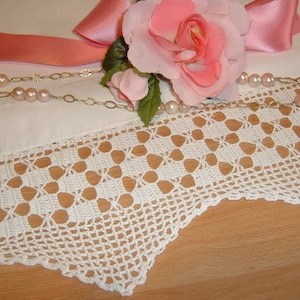 Może przedstawiać: Biała koronka haftowana na szydełku z falowanym brzegiem. Różowa satynowa wstążka z różową różą leży na białej powierzchni tkaniny. Naszyjnik z pereł jest udrapowany na tkaninie.