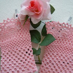 May include: Two pale pink, open-weave crochet wrist cuffs with a scalloped edge. The cuffs are displayed with a pink rose and green leaves against a white background. The cuffs are made of a delicate, lacy yarn.
