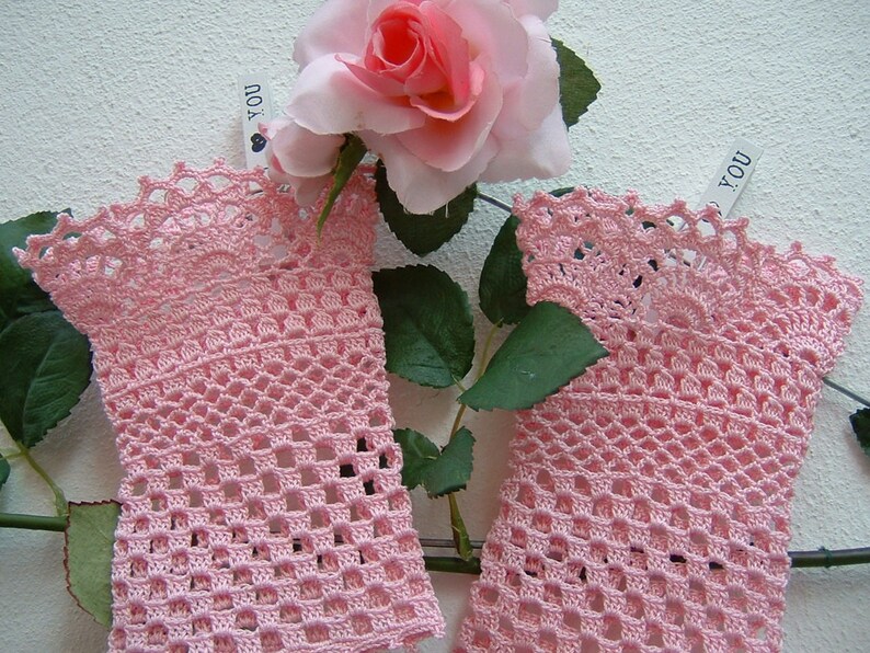 May include: Two pale pink, open-weave crochet wrist cuffs with a scalloped edge. The cuffs are displayed with a pink rose and green leaves against a white background. The cuffs are made of a delicate, lacy yarn.