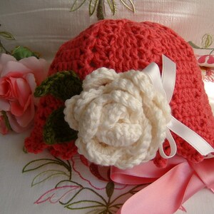 May include: A crocheted hat in a bright coral color with a white crocheted flower and a pink satin ribbon.