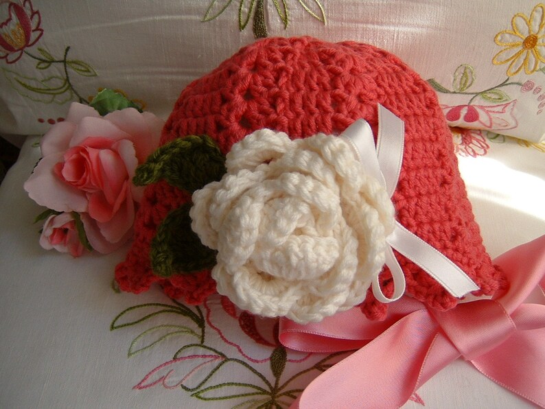 May include: A crocheted hat in a bright coral color with a white crocheted flower and a pink satin ribbon.