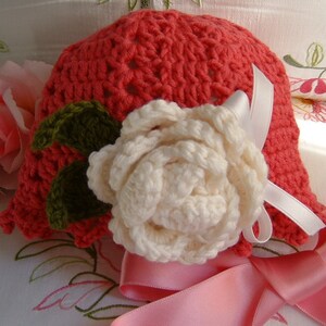May include: A crocheted hat in a bright coral color with a white crocheted flower and green leaves. The hat is decorated with a white satin ribbon.