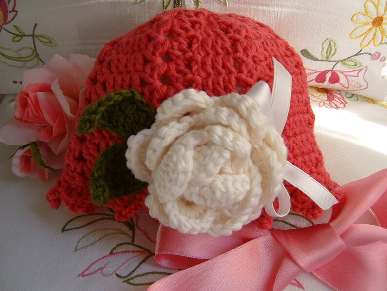 May include: A crocheted hat in a bright coral color with a white crocheted flower and green leaves. The hat is decorated with a white satin ribbon.