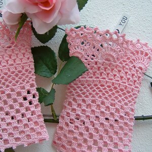 May include: Two pieces of delicate, light pink crocheted lace, possibly fingerless gloves or wrist cuffs, are displayed. The openwork design is detailed, with a scalloped edge. A pink rose and green leaves are in the background.