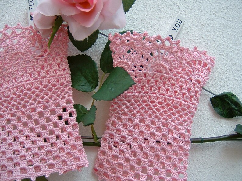 May include: Two pieces of delicate, light pink crocheted lace, possibly fingerless gloves or wrist cuffs, are displayed. The openwork design is detailed, with a scalloped edge. A pink rose and green leaves are in the background.