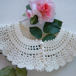 May include: A white crocheted collar with a pearl button in the center. The collar is displayed on a hanger with a pink rose behind it.