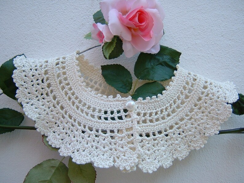 May include: A white crocheted collar with a pearl button in the center. The collar is displayed on a hanger with a pink rose behind it.