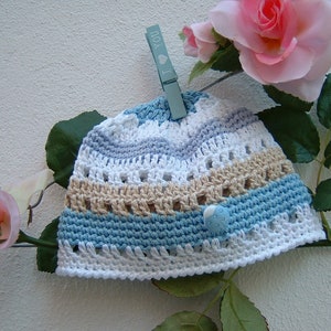 Könnte beinhalten: Eine gehäkelte Baby-Mütze mit weißen, blauen und beigen Streifen. Die Mütze hat einen blauen Knopf mit einem weißen Punkt oben. Die Mütze hängt an einer Wäscheleine mit einer blauen Wäscheklammer, auf der "Ich liebe dich" steht.