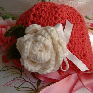 May include: A crocheted hat in a bright coral color with a white crocheted rose and a pink satin ribbon.