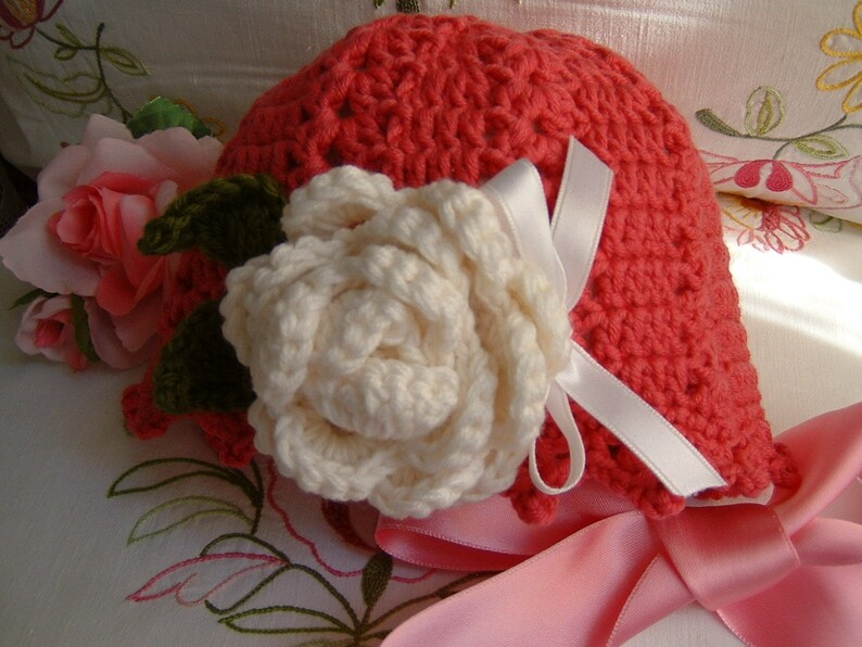May include: A crocheted hat in a bright coral color with a white crocheted rose and a pink satin ribbon.