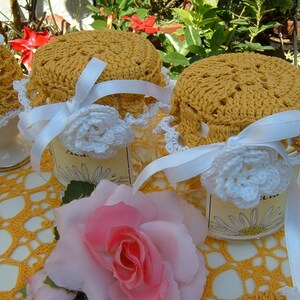 May include: Three crocheted jar covers in a yellow colour with white crocheted flowers and white ribbon bows. The covers are on top of glass jars with labels that have a daisy design. The jars are sitting on a white crocheted doily.