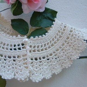 May include: A white crocheted collar with a pearl button in the center. The collar is on a hanger and has a scalloped edge.
