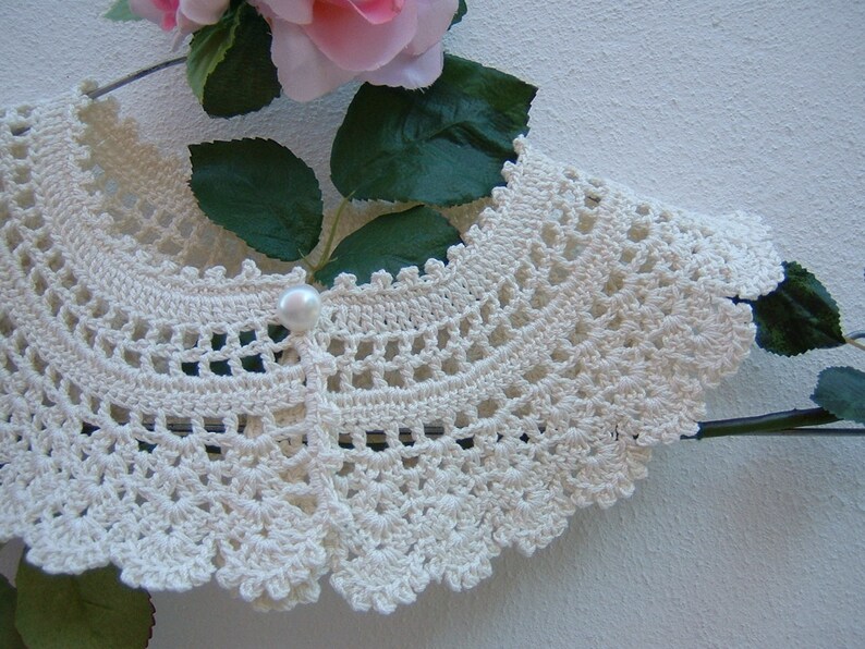 May include: A white crocheted collar with a pearl button in the center. The collar is on a hanger and has a scalloped edge.