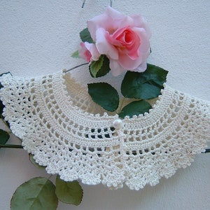 May include: A white crocheted collar with a pearl button in the center. The collar is displayed on a hanger with a pink rose and green leaves.