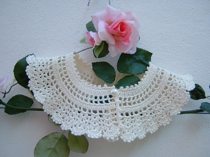 May include: A white crocheted collar with a pearl button in the center. The collar is displayed on a hanger with a pink rose and green leaves.