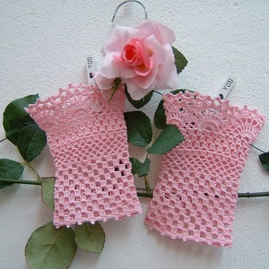 May include: Two pale pink, open-weave crochet gloves hang on a silver hanger, decorated with a pink rose and green leaves. The gloves feature a scalloped edge and a delicate, lacy design. A small tag with the word "YOU" is attached.