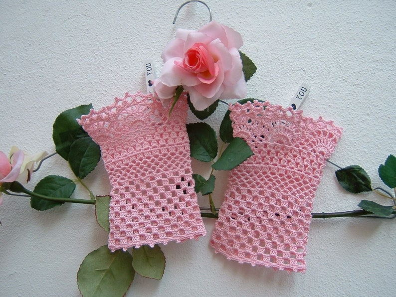 May include: Two pale pink, open-weave crochet gloves hang on a silver hanger, decorated with a pink rose and green leaves. The gloves feature a scalloped edge and a delicate, lacy design. A small tag with the word "YOU" is attached.