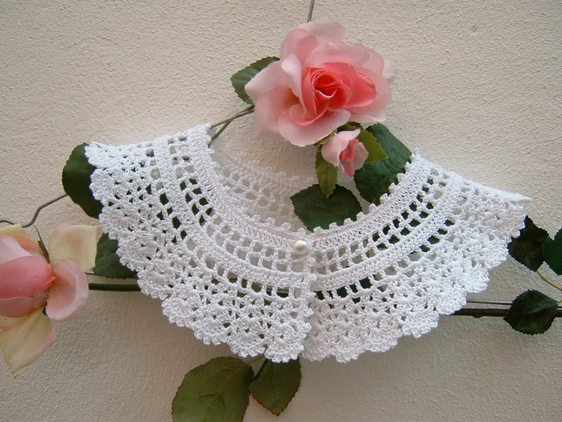 May include: White crocheted collar with a pearl button closure. The collar is on a hanger with pink roses and green leaves.
