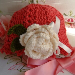 May include: A crocheted hat in coral pink with a white crocheted flower and green leaves. The hat is tied with a pink satin ribbon.