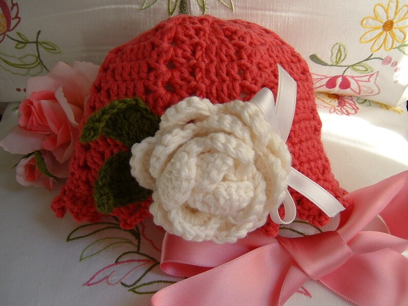 May include: A crocheted hat in coral pink with a white crocheted flower and green leaves. The hat is tied with a pink satin ribbon.