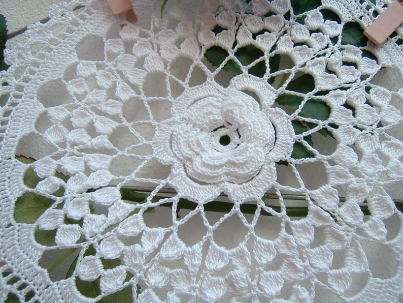 Puede incluir: Doily de ganchillo blanco con un delicado dise&ntilde;o floral. El doily presenta una gran flor en relieve en el centro, rodeada de motivos florales m&aacute;s peque&ntilde;os e intrincados.