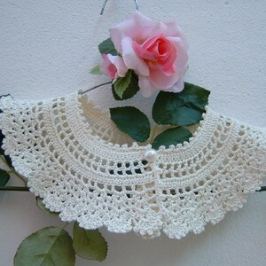 May include: A white crocheted collar with a pearl button in the center. The collar is displayed on a hanger with a pink rose.