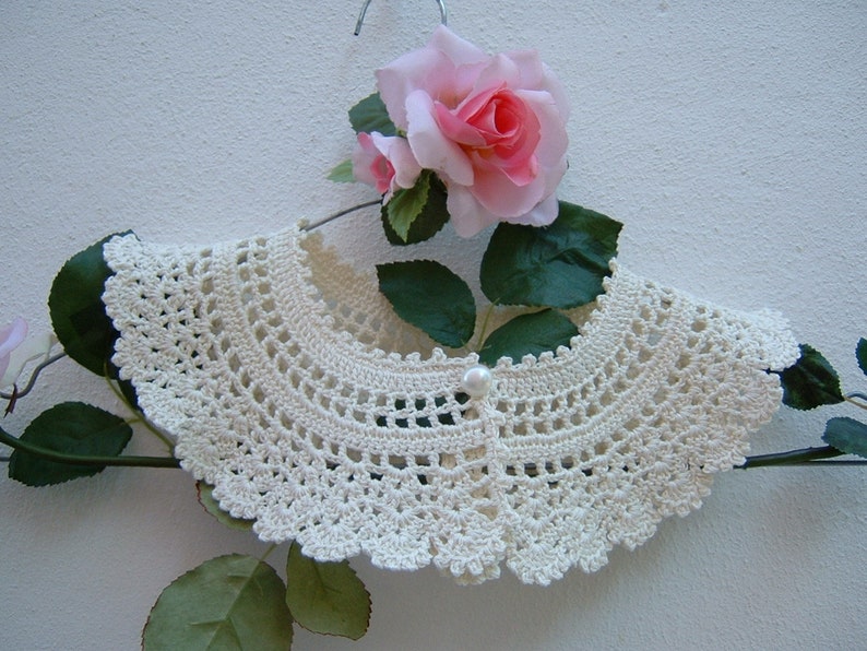 May include: A white crocheted collar with a pearl button in the center. The collar is displayed on a hanger with a pink rose.