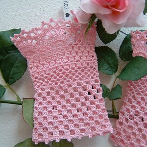 May include: Two light pink crochet arm warmers with an openwork design. The arm warmers are displayed with a pink rose and green leaves, creating a soft and feminine aesthetic. The crochet work features intricate patterns and a scalloped edge.