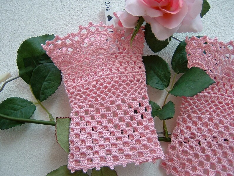 May include: Two light pink crochet arm warmers with an openwork design. The arm warmers are displayed with a pink rose and green leaves, creating a soft and feminine aesthetic. The crochet work features intricate patterns and a scalloped edge.