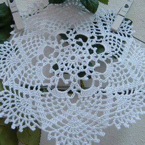 Peut inclure: Nappe blanche au crochet avec un motif floral. La nappe ronde pr&eacute;sente un motif ajour&eacute; complexe et un bord festonn&eacute;. Elle est maintenue par deux pinces &agrave; linge, avec des feuilles vertes et un mur blanc en arri&egrave;re-plan.