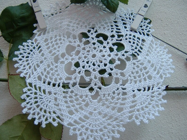 Peut inclure: Nappe blanche au crochet avec un motif floral. La nappe ronde pr&eacute;sente un motif ajour&eacute; complexe et un bord festonn&eacute;. Elle est maintenue par deux pinces &agrave; linge, avec des feuilles vertes et un mur blanc en arri&egrave;re-plan.