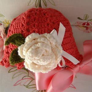 May include: A crocheted hat in a coral color with a white crocheted flower and green leaves. The hat has a pink satin ribbon tied around it.