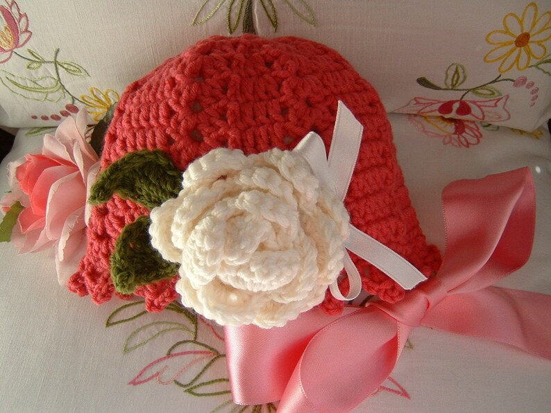 May include: A crocheted hat in a coral color with a white crocheted flower and green leaves. The hat has a pink satin ribbon tied around it.