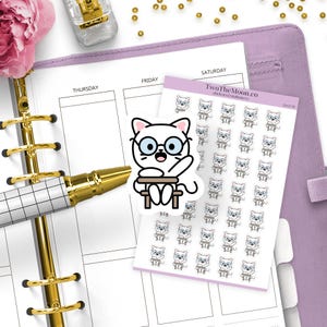 Puede incluir: Un planificador abierto con una pegatina de un gato blanco con gafas. La pegatina principal muestra un gato sentado en un escritorio. También hay una hoja de pegatinas con el mismo diseño de gato.