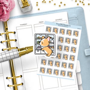Può includere: Un'agenda con una penna dorata, un fiore rosa e un foglio di adesivi con un cane corgi. Gli adesivi mostrano un cane corgi che apre un frigorifero. L'agenda ha una copertina azzurra e dettagli dorati.