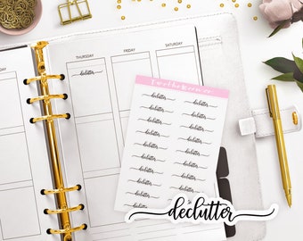May include: A planner with gold accents, a gold pen, and a pink sticker sheet with the word "declutter" in black script. The planner is open to a weekly spread, with a gold binder clip and scattered gold beads.