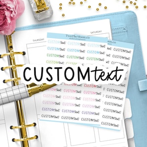 Op de afbeelding: Een vel met gepersonaliseerde stickers met de tekst CUSTOM text in verschillende kleuren, waaronder blauw, groen, roze, paars en zwart. De stickers liggen op een plannerpagina met een gouden ringbandmechanisme en een pen met ruitpatroon.