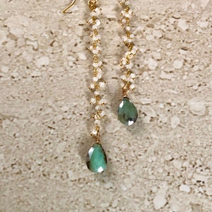 May include: A pair of long, gold dangle earrings with a teardrop-shaped green gemstone at the bottom of each earring. The earrings are made with a chain of small, white beads.