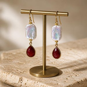 May include: A pair of gold-tone drop earrings featuring a rectangular iridescent white pearl suspended above a faceted teardrop-shaped deep red gemstone. The earrings hang from gold-tone hooks and include small gold-tone decorative bead accents.