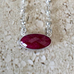 May include: A necklace featuring a horizontal oval-shaped, faceted deep red gemstone set in a silver bezel. The pendant is attached to a delicate silver chain that includes small, reflective, multifaceted silver beads spaced along the length.