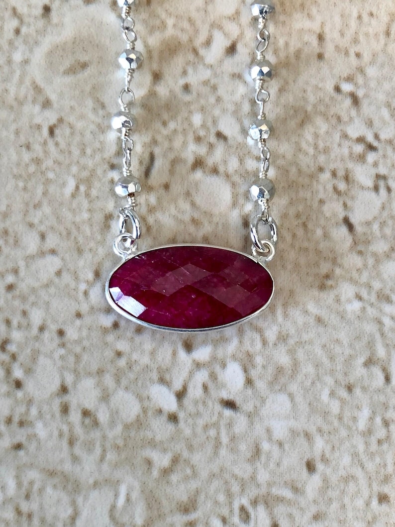 May include: A necklace featuring a horizontal oval-shaped, faceted deep red gemstone set in a silver bezel. The pendant is attached to a delicate silver chain that includes small, reflective, multifaceted silver beads spaced along the length.